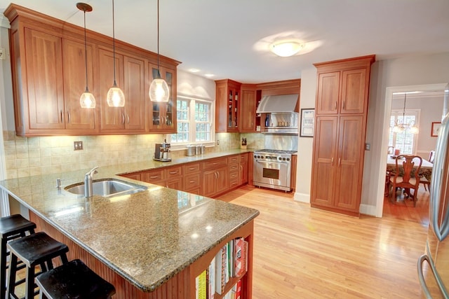 kitchen featuring a peninsula, wall chimney exhaust hood, brown cabinets, a sink, and appliances with stainless steel finishes