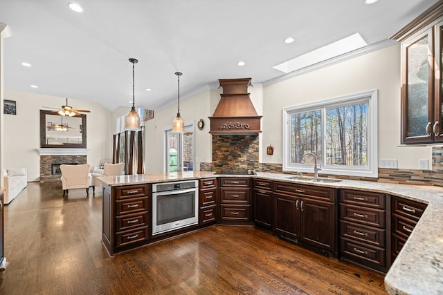 kitchen with custom exhaust hood, a sink, a stone fireplace, oven, and a peninsula