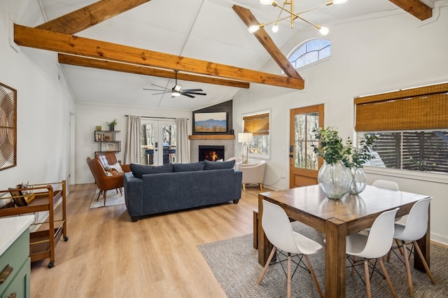 dining space featuring a warm lit fireplace, vaulted ceiling with beams, french doors, light wood-style floors, and ceiling fan with notable chandelier