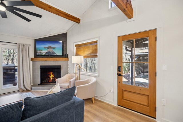 living area featuring light wood-type flooring, a warm lit fireplace, vaulted ceiling with beams, and ceiling fan