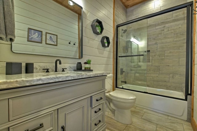 full bathroom with wood walls, bath / shower combo with glass door, vanity, and toilet