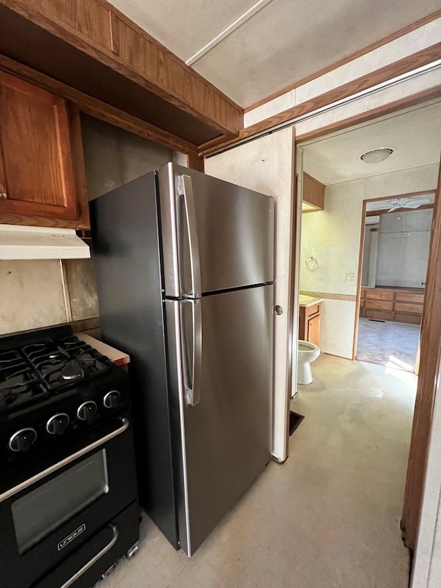 kitchen featuring concrete floors, under cabinet range hood, black gas range, freestanding refrigerator, and brown cabinetry