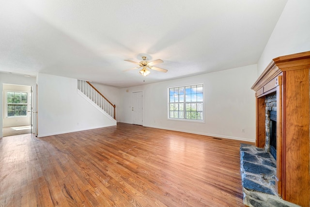 unfurnished living room with ceiling fan, a stone fireplace, baseboards, stairs, and light wood-style floors