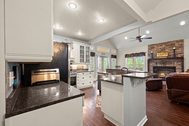 kitchen with dark stone counters, a stone fireplace, an island with sink, a kitchen bar, and white cabinetry