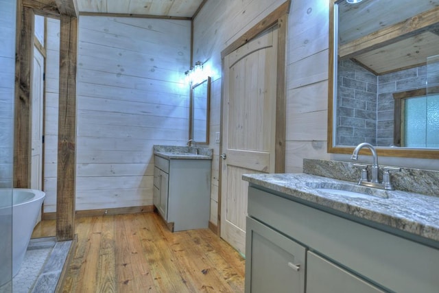 full bath with wooden walls, a sink, two vanities, a freestanding tub, and hardwood / wood-style flooring