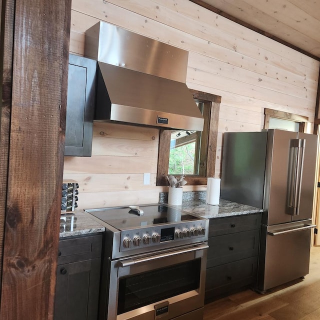 kitchen featuring stainless steel appliances, dark cabinets, wall chimney range hood, light stone countertops, and wooden walls