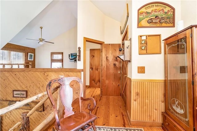 interior space with wooden walls, a wainscoted wall, high vaulted ceiling, ceiling fan, and light wood-type flooring