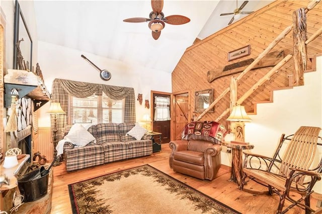 living room with stairway, wooden walls, wood finished floors, high vaulted ceiling, and ceiling fan