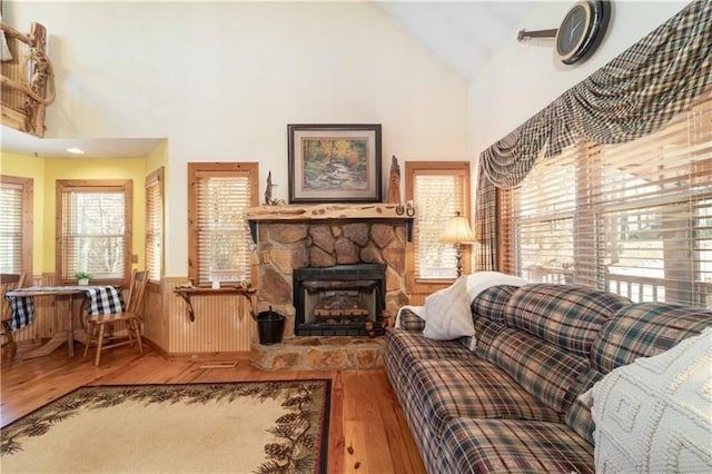 living area with a stone fireplace, wood finished floors, a wainscoted wall, and high vaulted ceiling