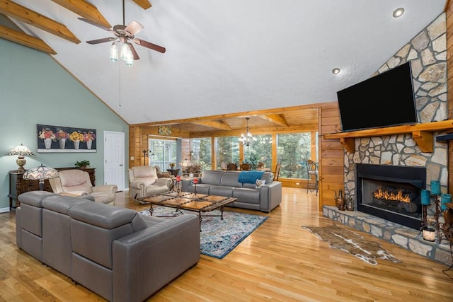 living area with ceiling fan with notable chandelier, high vaulted ceiling, wood finished floors, and a stone fireplace