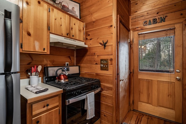 kitchen featuring brown cabinets, stainless steel appliances, light countertops, wood walls, and under cabinet range hood