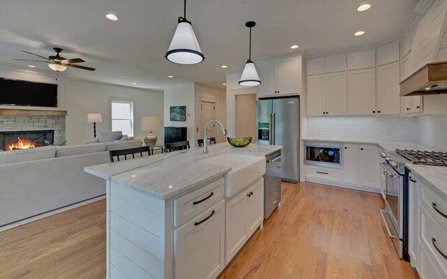 kitchen with appliances with stainless steel finishes, white cabinets, a fireplace, hanging light fixtures, and an island with sink