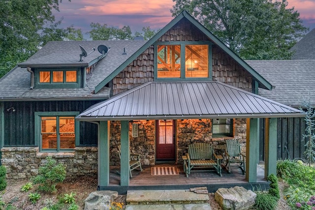 rear view of house with stone siding, a shingled roof, board and batten siding, metal roof, and a porch
