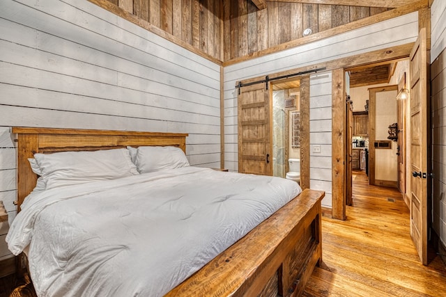 bedroom with wooden walls, a barn door, light wood-style flooring, ensuite bathroom, and vaulted ceiling