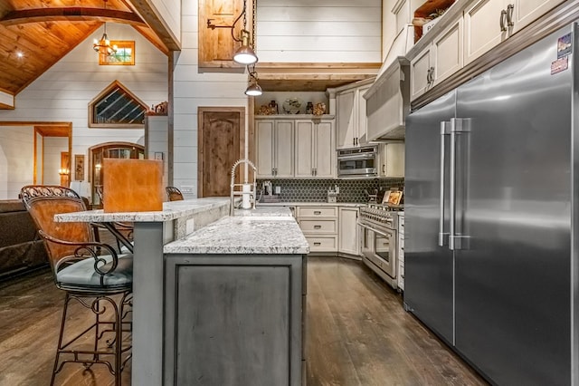 kitchen with light stone counters, premium appliances, a kitchen island with sink, pendant lighting, and dark hardwood / wood-style floors