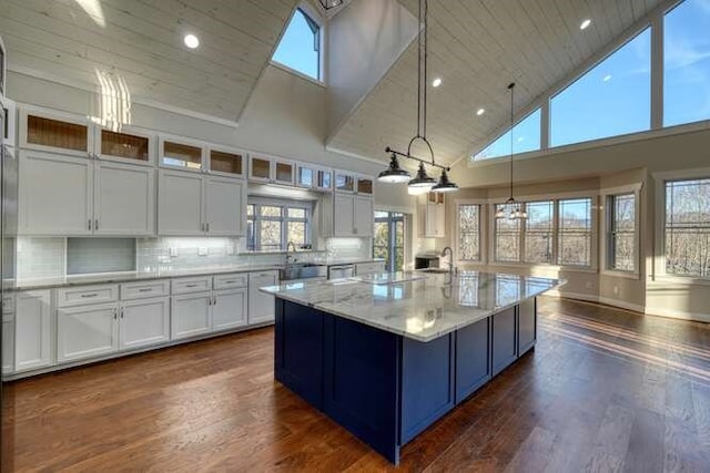 kitchen with high vaulted ceiling, tasteful backsplash, white cabinetry, light stone counters, and a center island with sink