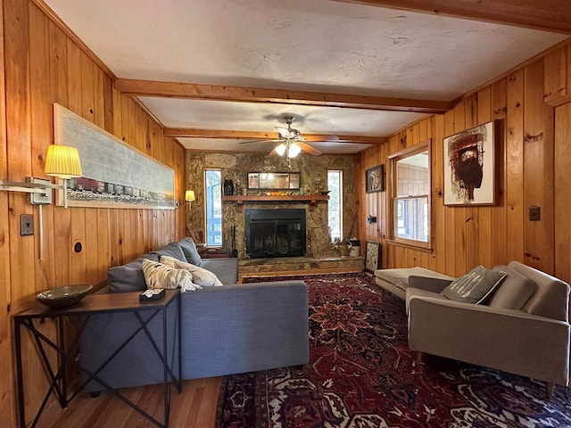 living area featuring wooden walls, wood finished floors, beamed ceiling, a fireplace, and ceiling fan