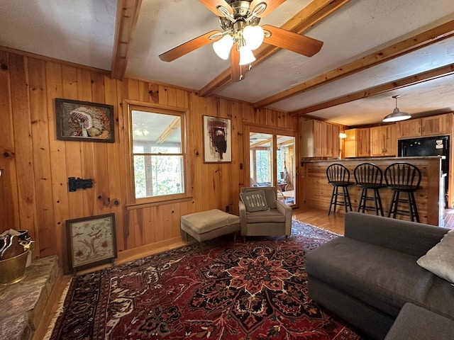 living area with ceiling fan, wood finished floors, beam ceiling, and wooden walls
