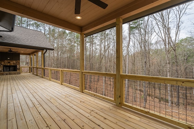 deck featuring an outdoor stone fireplace, a ceiling fan, and a view of trees