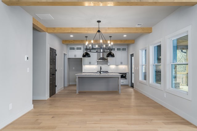 kitchen featuring wall chimney range hood, white cabinets, a chandelier, baseboards, and beamed ceiling