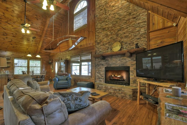 living room with wooden ceiling, wood walls, a stone fireplace, wood-type flooring, and ceiling fan