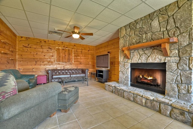 living room with wood walls, a stone fireplace, ceiling fan, and a drop ceiling