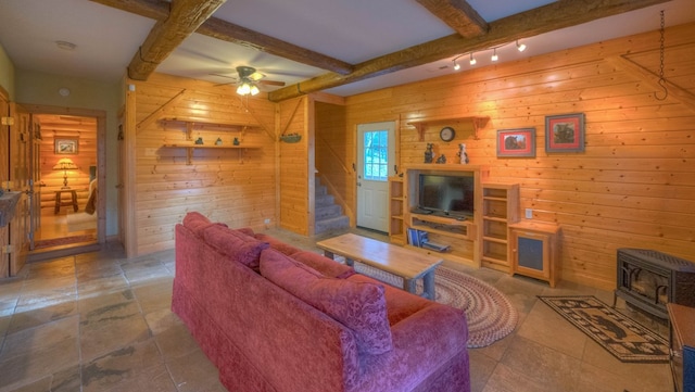 living room featuring wooden walls, a wood stove, ceiling fan, and beam ceiling