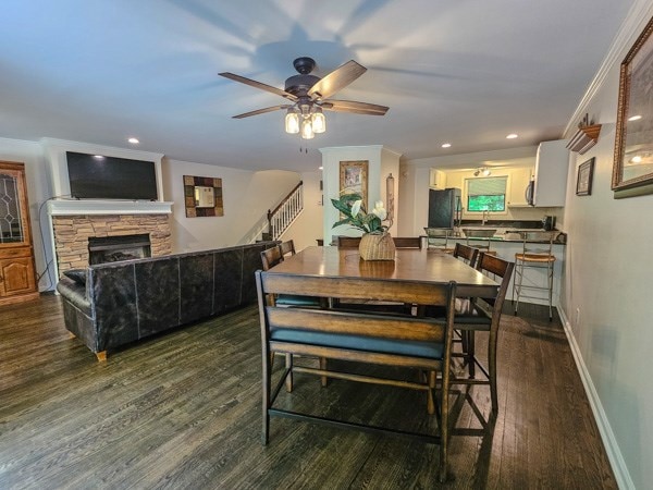 dining space featuring stairs, dark wood-style flooring, a stone fireplace, baseboards, and a ceiling fan