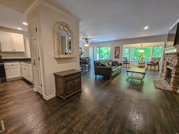 living area featuring a ceiling fan, a stone fireplace, ornamental molding, recessed lighting, and dark wood-type flooring