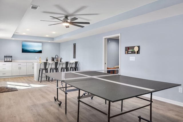 playroom with light wood-style flooring, bar with sink, a tray ceiling, a ceiling fan, and recessed lighting