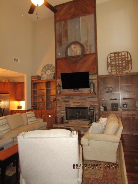 living room with ceiling fan, hardwood / wood-style flooring, a towering ceiling, and a stone fireplace