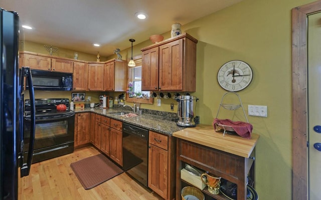 kitchen with a sink, light wood-style floors, hanging light fixtures, black appliances, and dark stone countertops