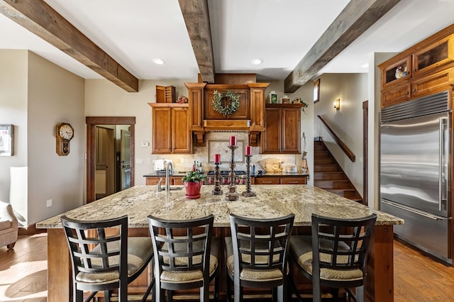 kitchen with beamed ceiling, tasteful backsplash, wood finished floors, built in fridge, and a large island with sink