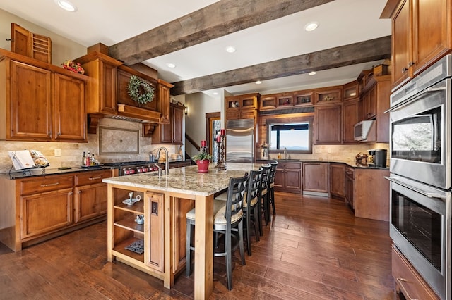 kitchen featuring beam ceiling, stainless steel appliances, dark wood-type flooring, and a kitchen island with sink