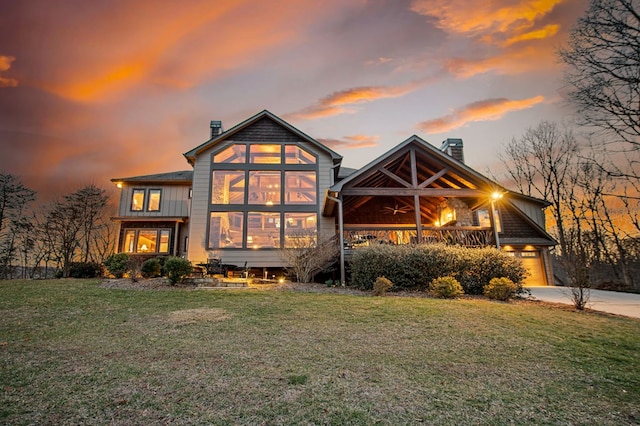 back of house at dusk with concrete driveway, a chimney, a yard, an attached garage, and a ceiling fan