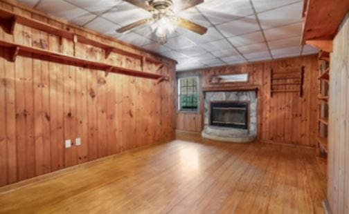 unfurnished living room with wooden walls, a drop ceiling, ceiling fan, wood finished floors, and a fireplace