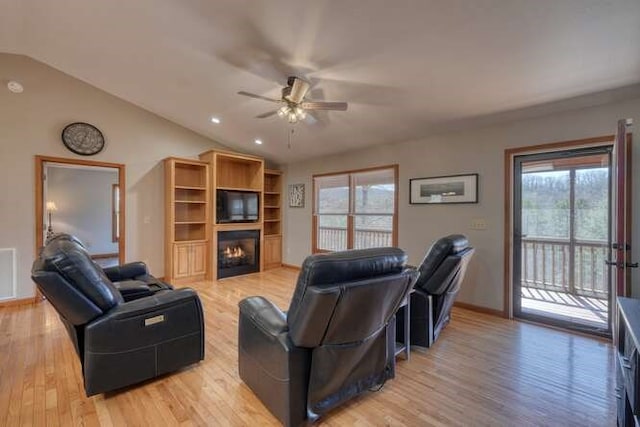 living area featuring lofted ceiling, visible vents, a ceiling fan, a glass covered fireplace, and light wood-type flooring