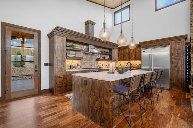 kitchen with dark wood-type flooring, wall chimney range hood, decorative light fixtures, built in refrigerator, and open shelves