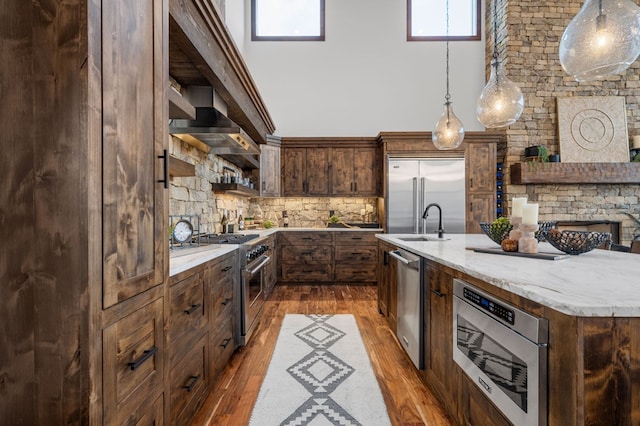 kitchen with an island with sink, decorative light fixtures, premium appliances, wall chimney range hood, and dark wood-style flooring