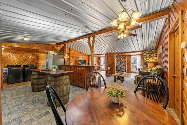 dining room featuring ceiling fan, lofted ceiling with beams, washer and dryer, a stone fireplace, and wood walls