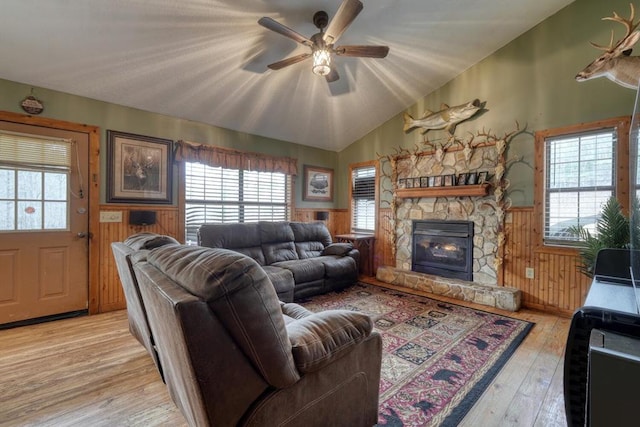 living area with light wood finished floors, a ceiling fan, wainscoting, vaulted ceiling, and a stone fireplace