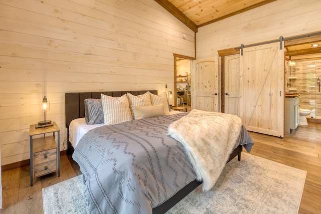 bedroom featuring wood walls, light hardwood / wood-style flooring, vaulted ceiling, a barn door, and connected bathroom