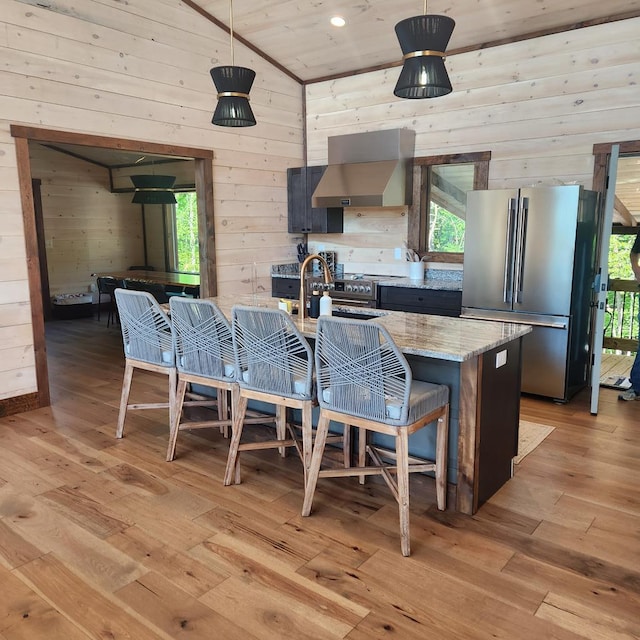 kitchen with appliances with stainless steel finishes, wall chimney exhaust hood, wooden walls, light hardwood / wood-style flooring, and lofted ceiling