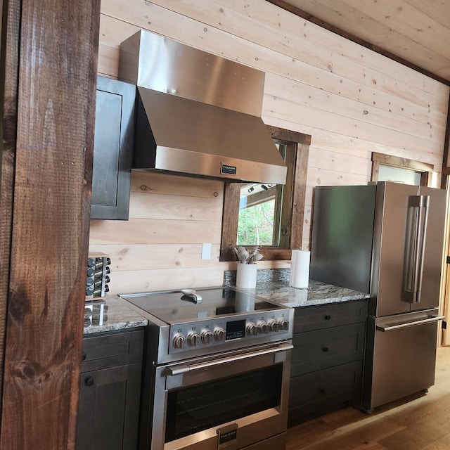 kitchen featuring wood walls, ventilation hood, light wood-type flooring, light stone countertops, and appliances with stainless steel finishes