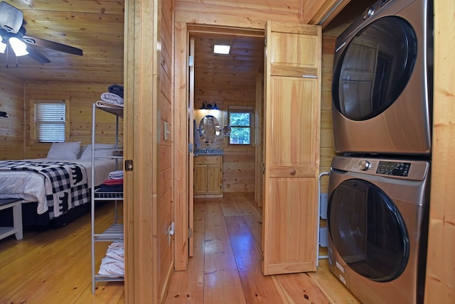 clothes washing area featuring a ceiling fan, wooden walls, light wood finished floors, stacked washer / dryer, and laundry area