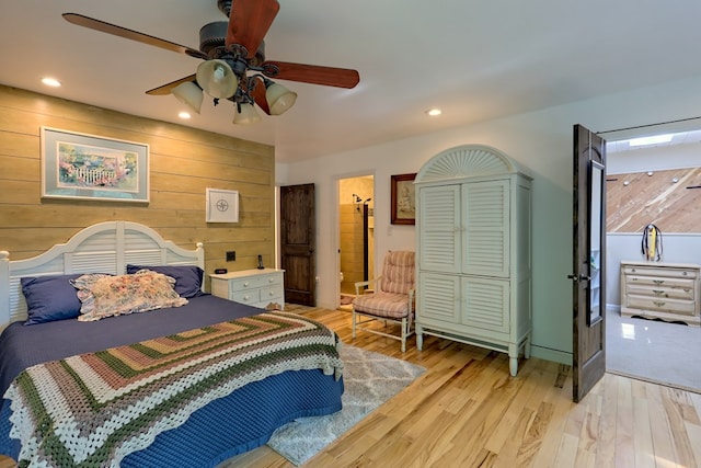 bedroom with ensuite bathroom, light hardwood / wood-style flooring, ceiling fan, and wood walls