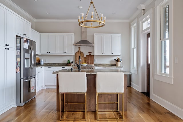 kitchen with stainless steel refrigerator with ice dispenser, wall chimney exhaust hood, a kitchen island with sink, white cabinetry, and a kitchen bar