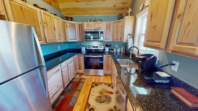 kitchen with light brown cabinetry, a sink, dark stone countertops, vaulted ceiling, and stainless steel appliances