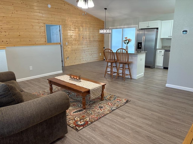 living area featuring a notable chandelier, light wood finished floors, wood walls, baseboards, and vaulted ceiling