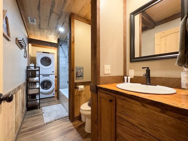 full bathroom featuring wood finished floors, a wainscoted wall, visible vents, stacked washer and clothes dryer, and toilet